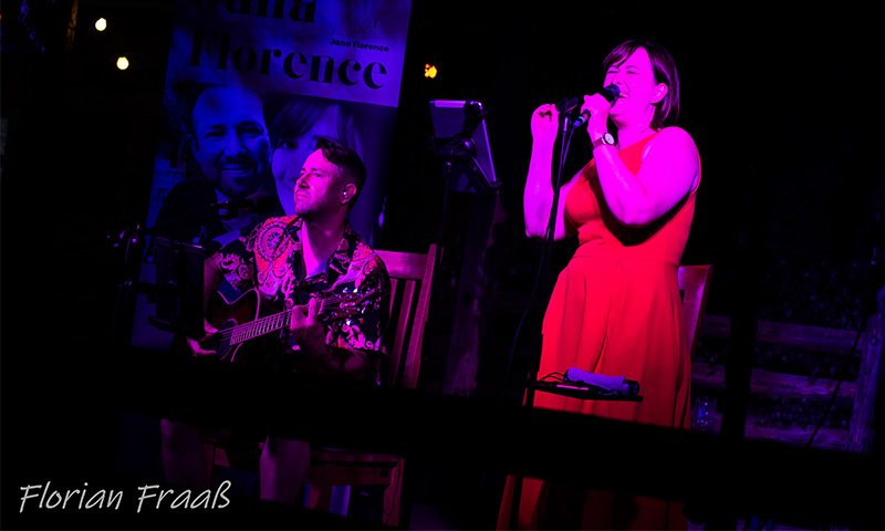 Jana in rotem Kleid mit Mikrofon in der Hand emotional singend und Danny mit Gitarre vor eigenem Jana Florence Roll-Up in pinkem Licht