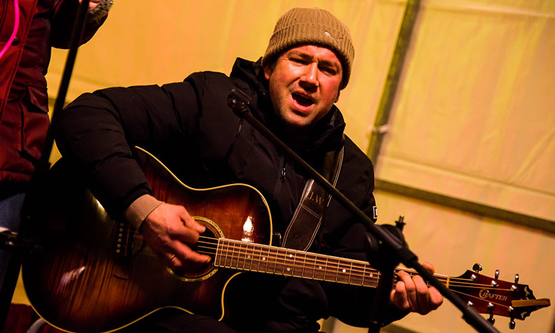 Danny mit brauner Mütze hinter einem Mikrofon am Gitarre spielen und singen