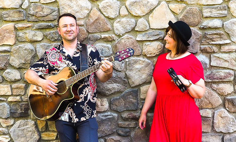 Jana und Danny vor einer Steinwand in Kleid und Hemd mit Gitarre und einem Shaker