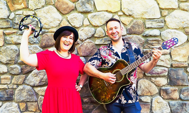 Jana und Danny lachend vor einer Steinwand in Kleid und Hemd mit Gitarre und einem Shaker, die Hände in die Luft streckend