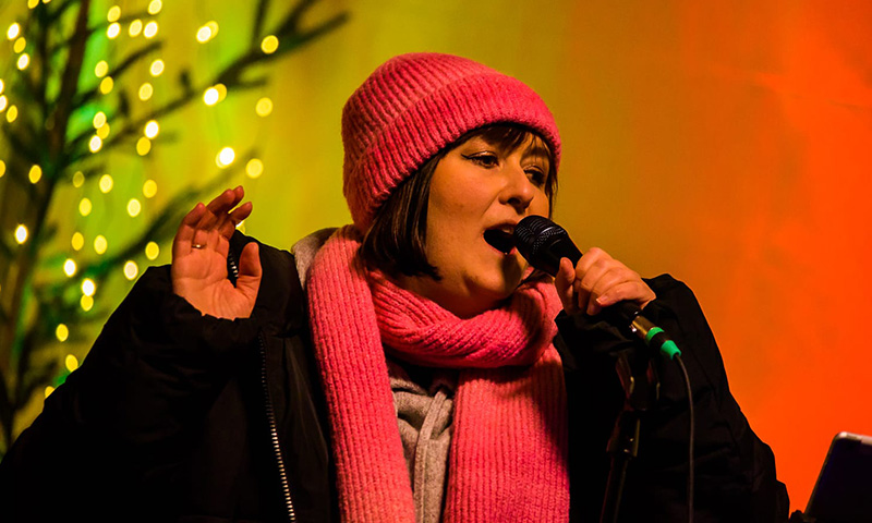 Jana in pinker Mütze und pinkem Schal mit Mikrofon in der Hand singend vor einem im Hintergrund unscharfen beleuchteten Baum